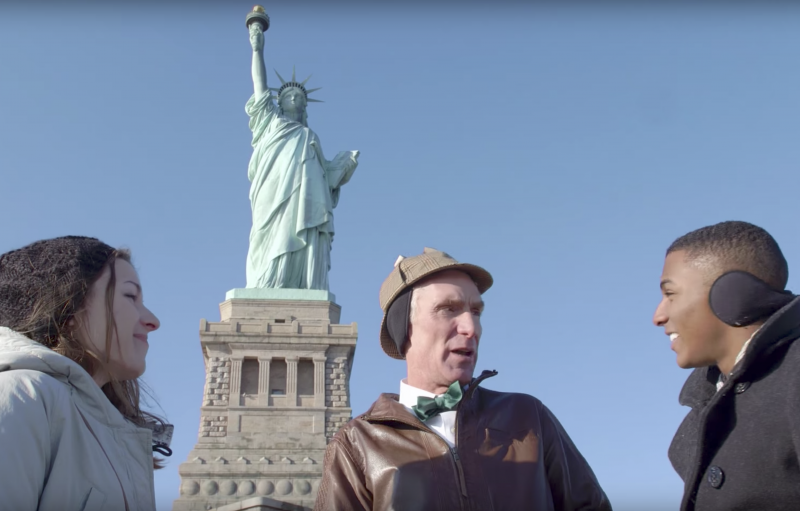 Bill Nye with two other people standing in front of the Statue of Liberty