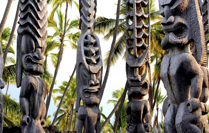 Cuatro tótems de madera iluminados por el sol con caras talladas en el Parque Nacional Pu'uhonua