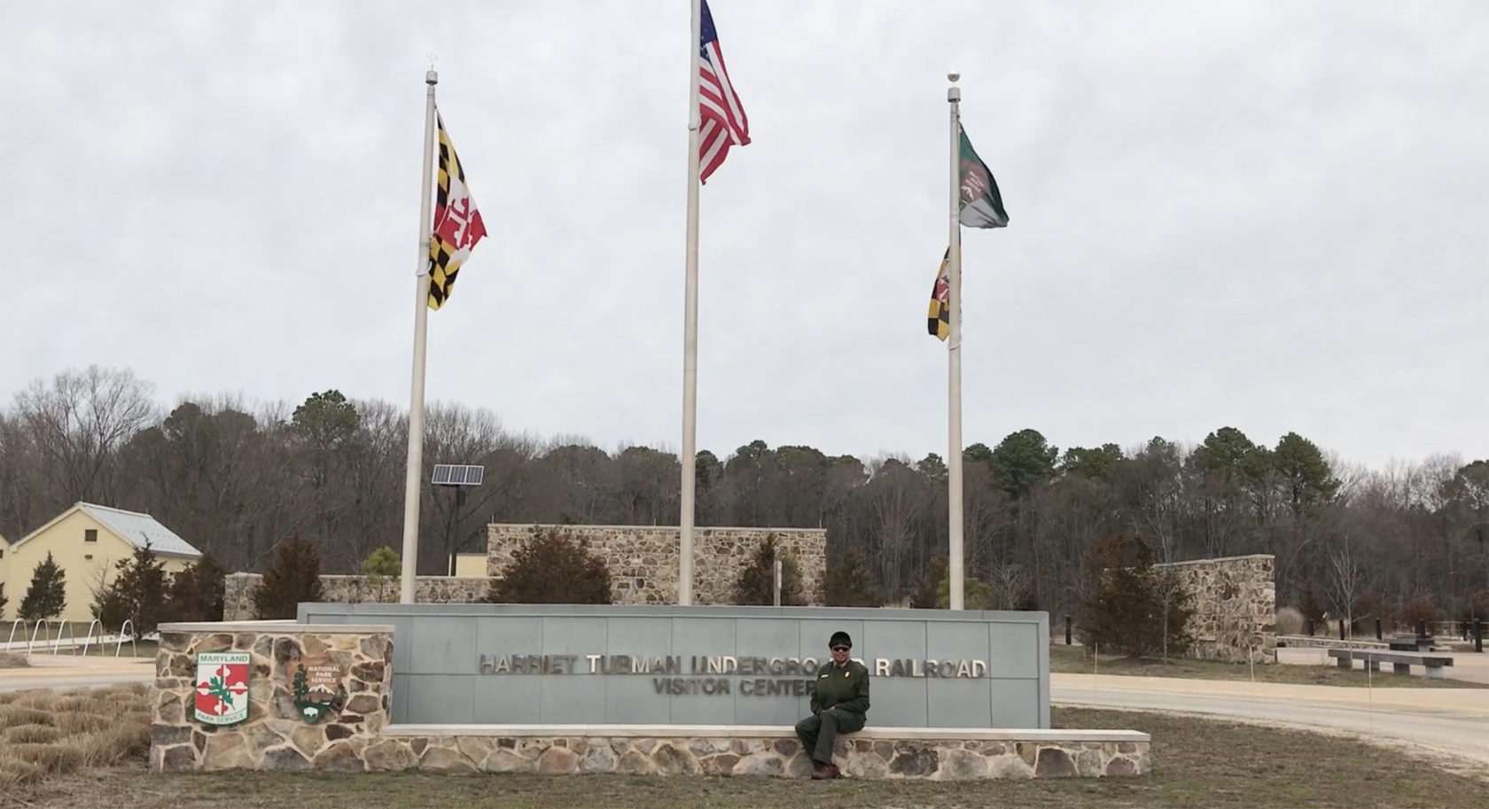 Deanna Mitchell sits outside on a stone sign for the Visitor Center at Harriet Tubman Underground Railroad National Historical Park. Three flags wave gently on flagpoles behind the sign.