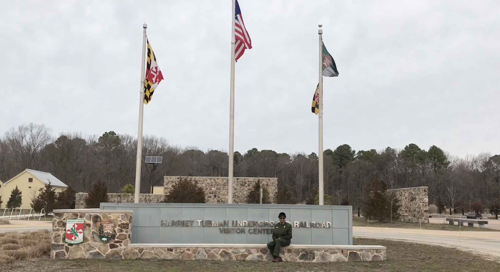 Deanna Mitchell se sienta afuera en un letrero de piedra para el Centro de Visitantes en el Parque Histórico Nacional del Ferrocarril Subterráneo Harriet Tubman. Tres banderas ondean suavemente en mástiles detrás del letrero.