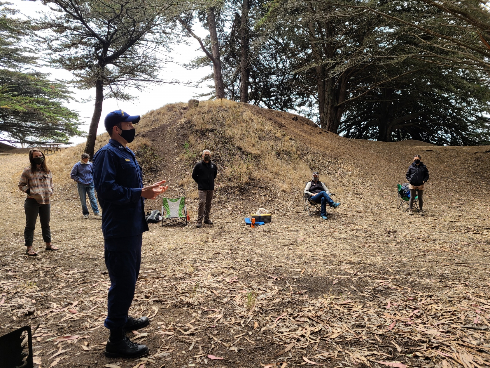 LCDR Matt Kozler, in a mask, talks to a group of people, also wearing masks and standing far away from each other