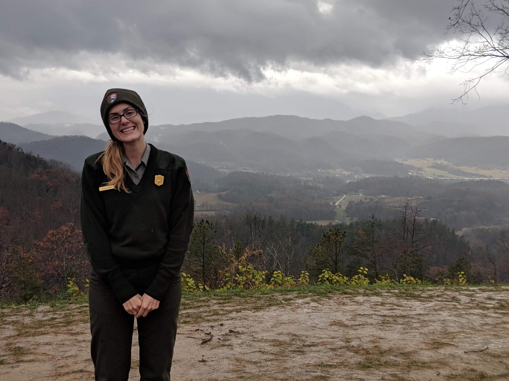 Jessie Snow, con uniforme de NPS, sonríe a la cámara. Ella se para frente a una vista de las Grandes Montañas Humeantes
