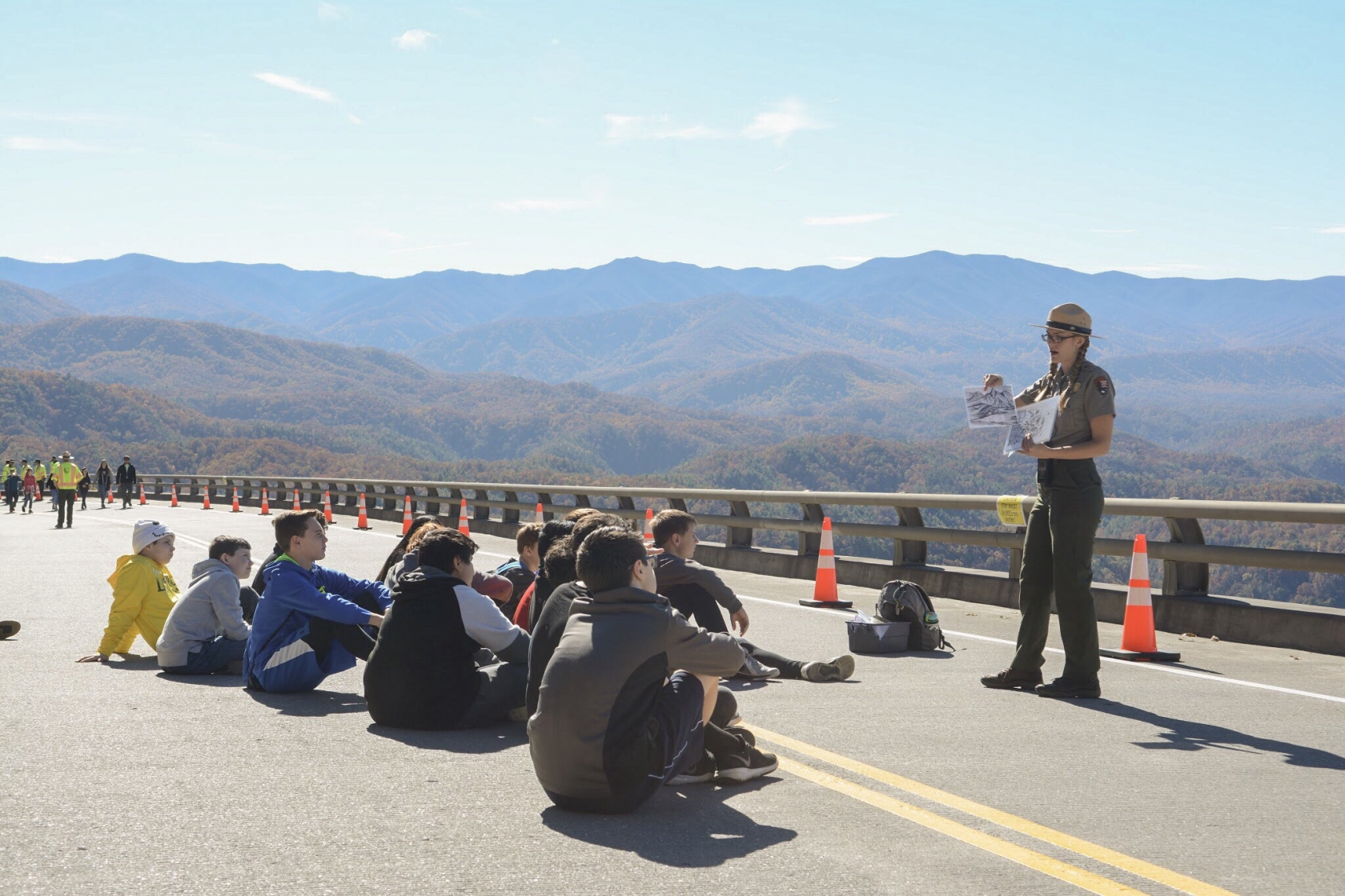 On a paved road, blocked off to traffic, Jessie Snow, in full NPS uniform, holds up a couple of papers to give a demonstration to a group of students sitting on the road in front of her. In the background, a scenic vista of the Great Smokies