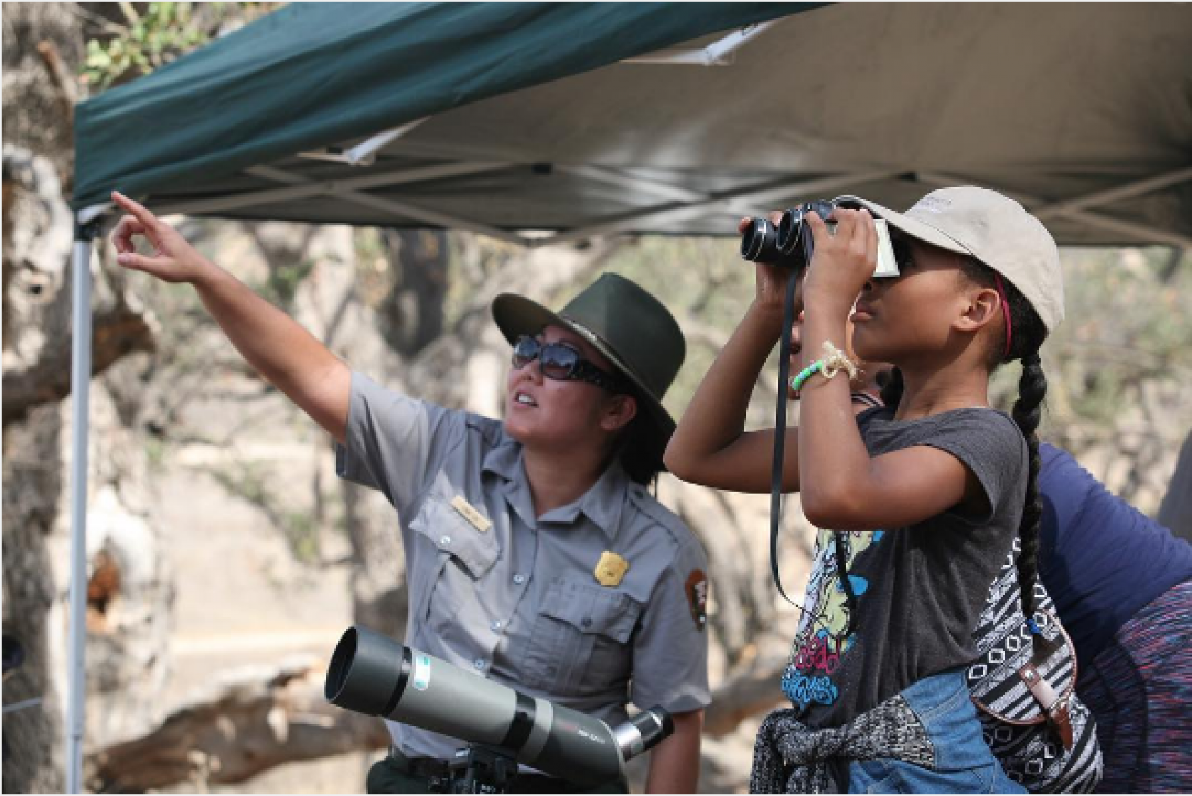 Park ranger pointing to sky while child looks through binoculars 
