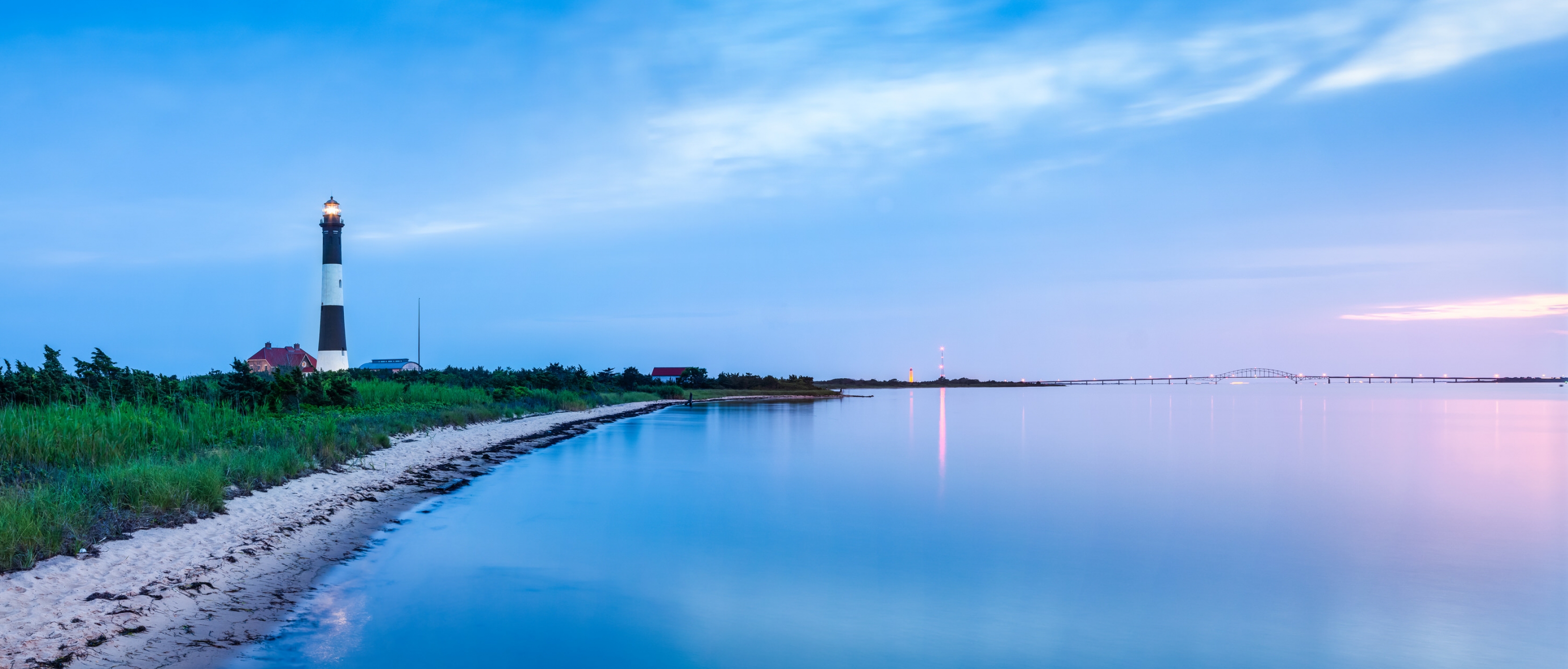 Blue dusk reflected into the ocean at the shore with a black and white lighthouse among greenery at Fire Islands National Seashore