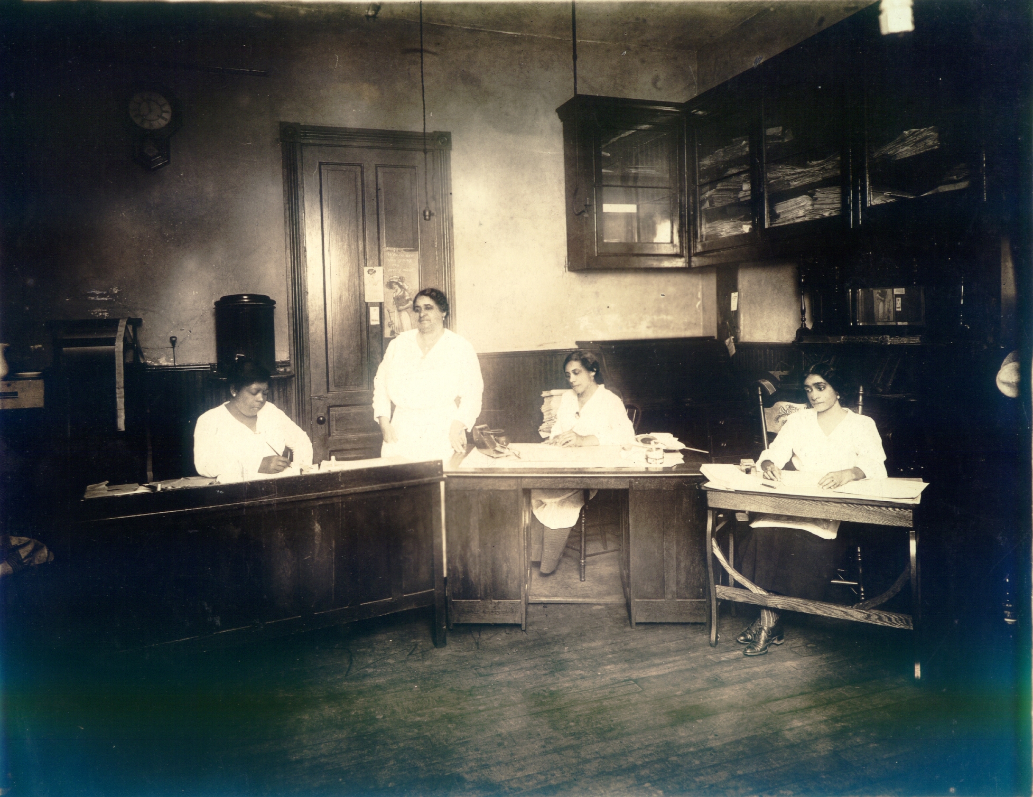 Una histórica fotografía en blanco y negro de tres mujeres sentadas en sus escritorios en la oficina del periódico St. Luke Herald con Maggie Walker de pie detrás de ellas.