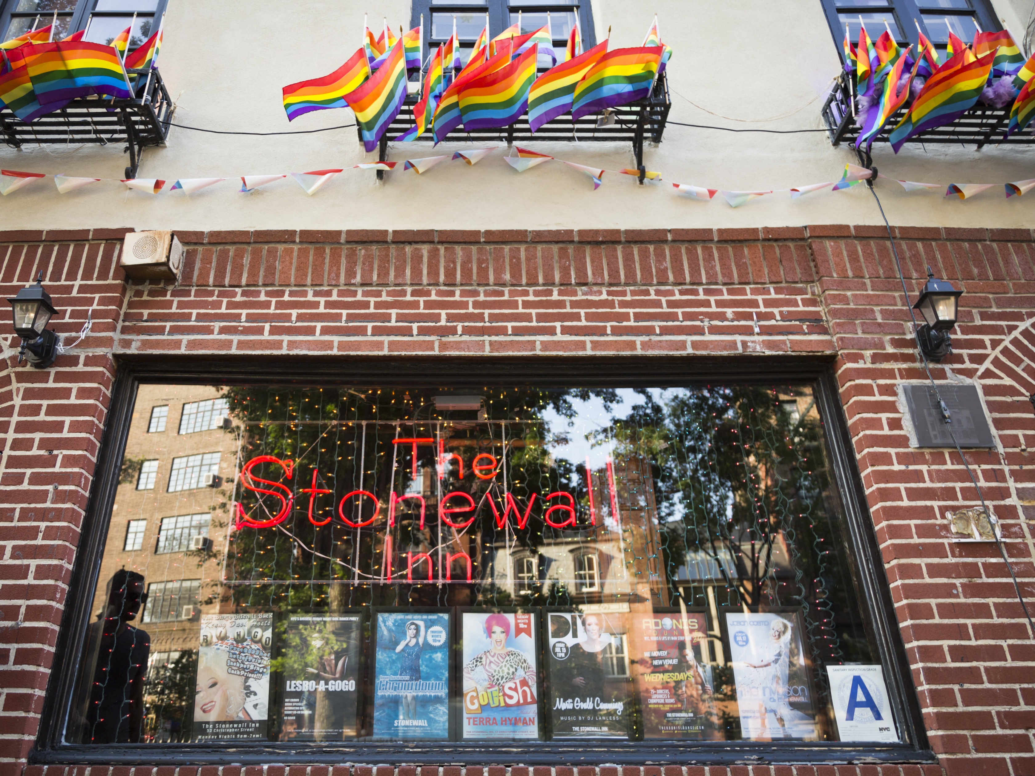 Neone sign saying "The Stonewall Inn" in a window of a brick building with many small rainbow flags flying above the window.