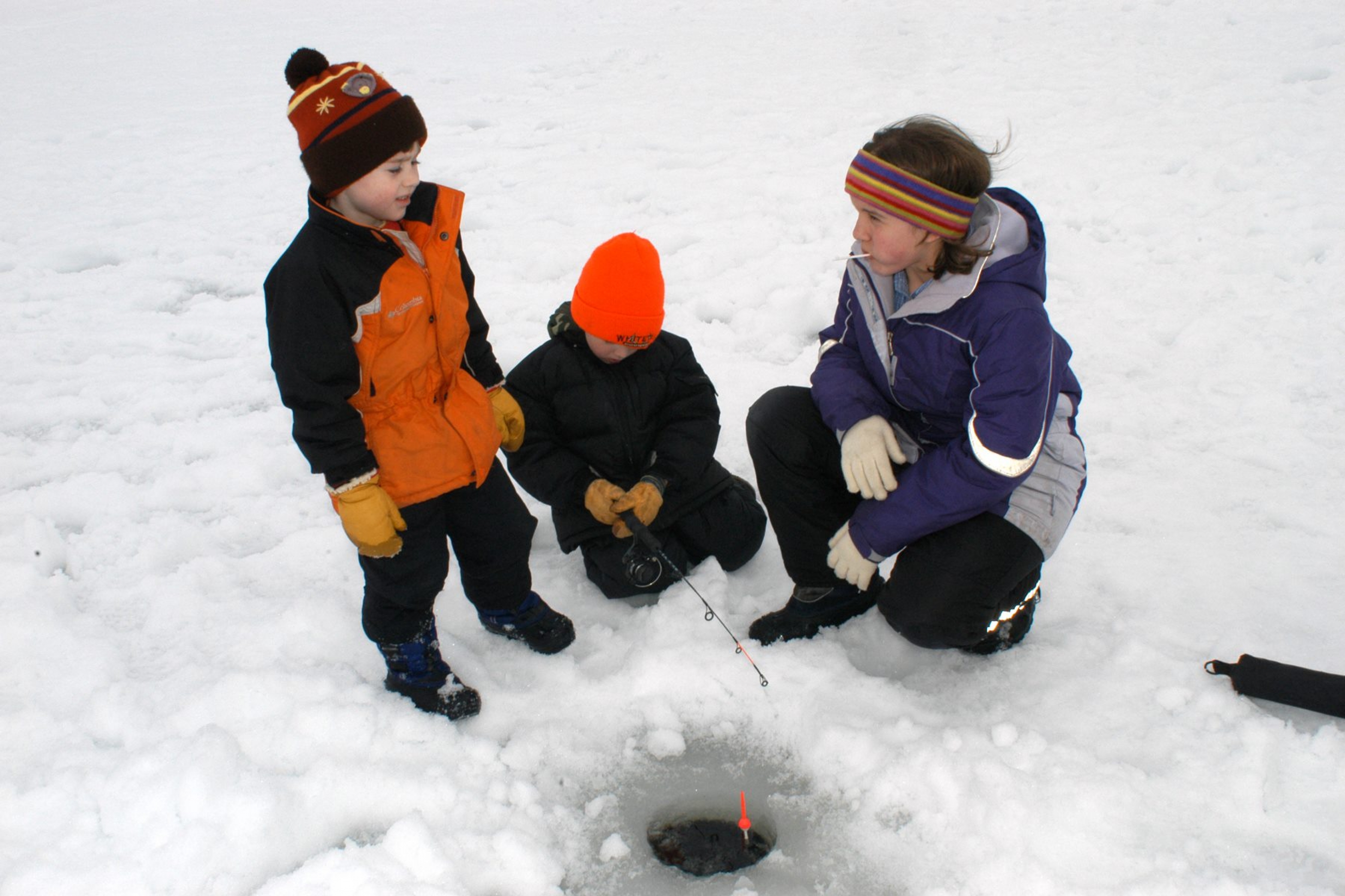 3 children on a snow-covered ice with a fishing rod through a small hole in the ice at Voyageurs National Park