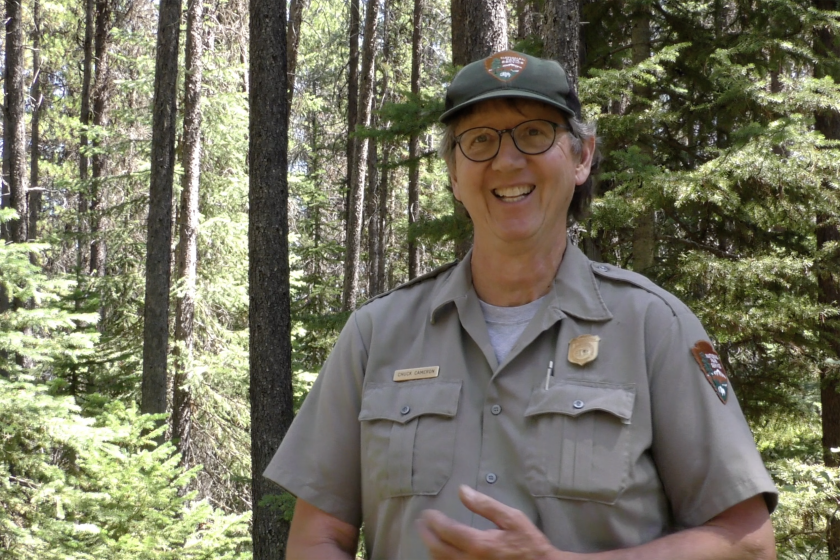 Ranger Chuck Cameron smiles at a camera, surrounded by green trees at Glacier National Park