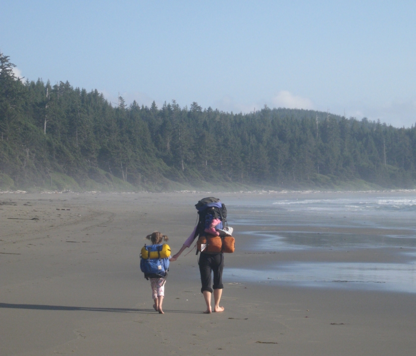 Child and adult with backpacks walking down a beach next to a forest at Olympic National Park